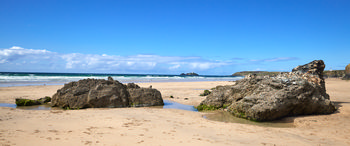 Gwithian rocks This landscape photograph shows the Gwithian rocks situated on a sandy beach in Cornwall, England, United Kingdom. Taken in the afternoon during the summer season, the image captures clear blue skies over the coastline and calm sea waves approaching the shore. In the distance, Godrevy Lighthouse is visible, which is a notable landmark along the Cornish coast. The beach scene highlights the natural rock formations and patches of wet sand, characteristic of the coastal area in England.